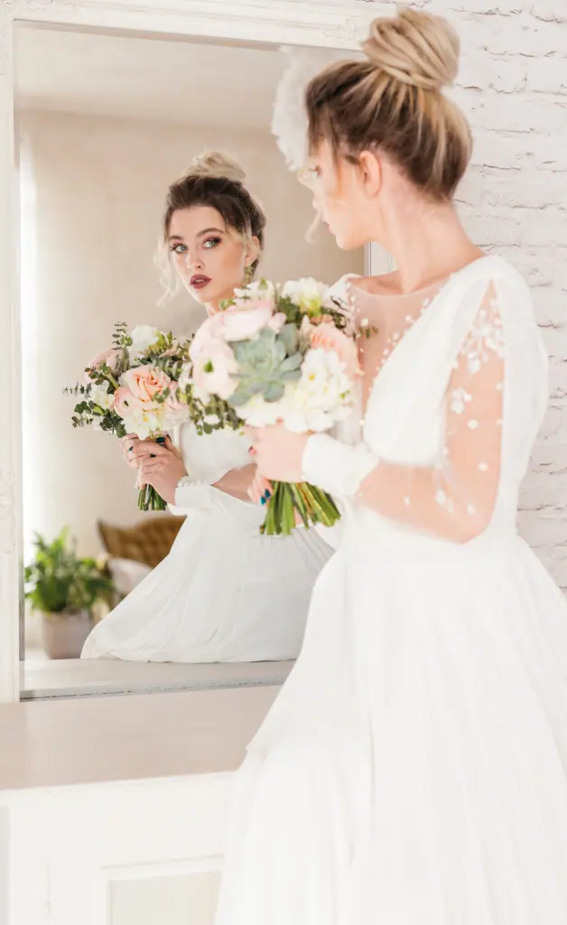 A girl wearing a white dress stands in front of a mirror, holding a bouquet of flowers, admiring her reflection.