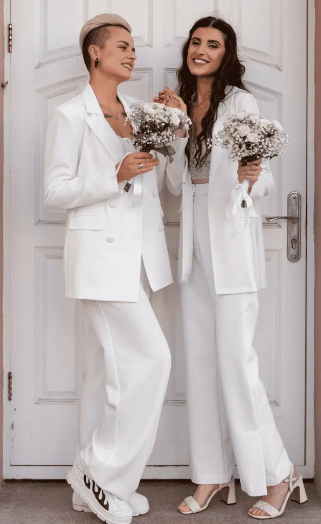 Mother and daughter, both wearing white dresses, holding a bouquet of flowers in their hands, standing together gracefully.