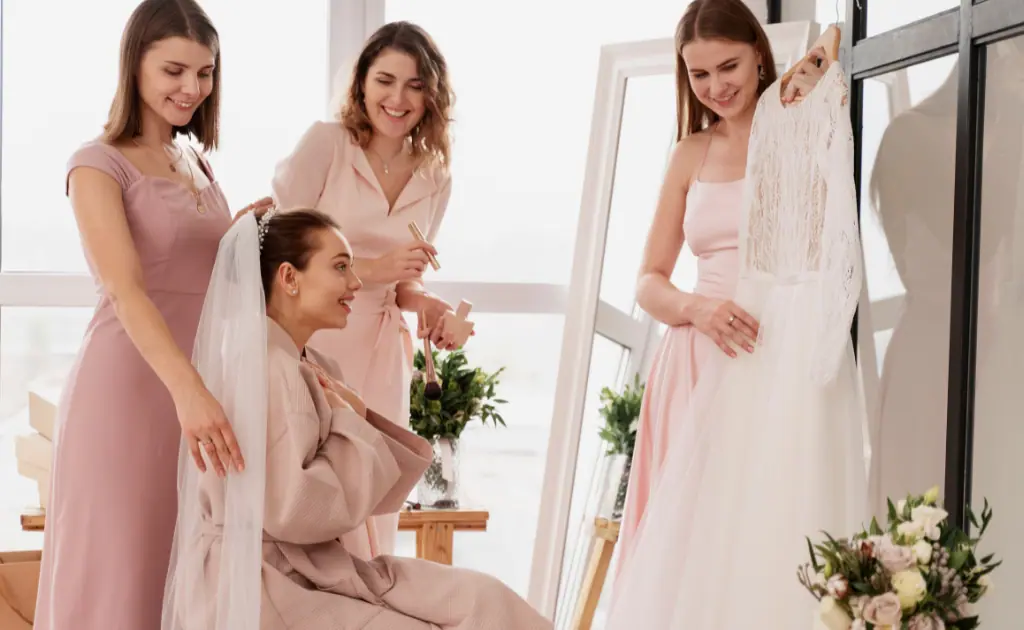 Three girls standing, showing a Mother of the Groom dress to a seated girl, who smiles joyfully as they all admire the dress together.